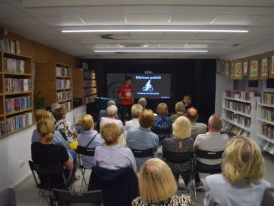 Szerokie ujęcie sali bibliotecznej z perspektywy tyłu widowni. Daniel Kocuj stoi na tle czarnej ściany obok dużego ekranu wyświetlającego napis „Bike’owa podróż z Sydney do Szczecina” z grafiką rowerzysty. Autor, ubrany w czerwoną koszulkę z godłem Polski, przemawia do zgromadzonej w rzędach krzeseł publiczności. Z obu stron salę otaczają wysokie regały pełne książek