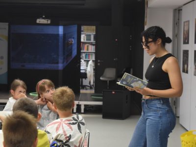 Prowadząca w okularach i czarnym topie stoi w sali bibliotecznej, czytając dzieciom trzymaną w rękach małą książeczkę. Przed nią siedzi grupa zasłuchanych dzieci