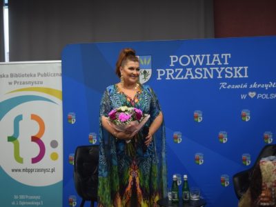 Katarzyna Skrzynecka w kolorowej, wzorzystej sukni trzyma bukiet kwiatów i uśmiecha się do publiczności na tle niebieskiej ścianki promocyjnej Powiatu Przasnyskiego i Miejskiej Biblioteki Publicznej w Przasnyszu