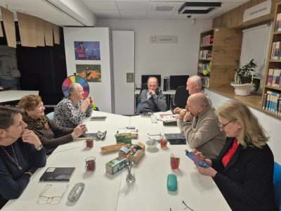 Siedem dorosłych osób zajmuje miejsca wokół stołu podczas spotkania Dyskusyjnego Klubu Książki. Część uczestników słucha, inni zaglądają do telefonów, a w tle widoczne są regały biblioteczne.