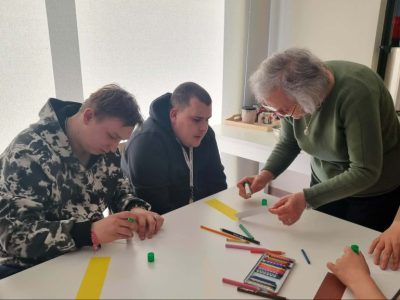 Trzy osoby siedzą przy białym stole przy oknie. Dwóch młodych mężczyzn jest skupionych na pracy nad projektami. Po prawej stronie starsza kobieta w zielonym swetrze (instruktorka) aktywnie pomaga jednemu z nich, trzymając mały przedmiot. Na stole rozłożone są materiały: kolorowe kartki, kredki i pastele w metalowym pudełku