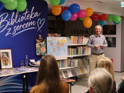 Uśmiechnięty Andrzej Ciuba stoi na wprost widowni na tle regału z mapą świata i kolorowych balonów pod sufitem. Na stole przed nim widoczne są oprawione zdjęcia i bibeloty z wypraw