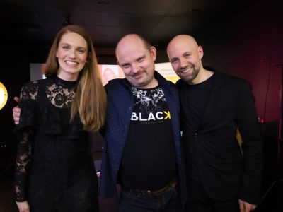 Natalia Srokocz i Jerzy Grzechnik pozują do zdjęcia, obejmując uśmiechniętego mężczyznę w koszuli i czarnym t-shircie. Wszyscy patrzą bezpośrednio w obiektyw