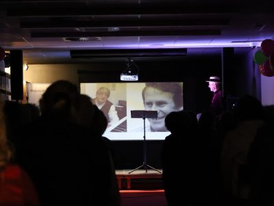 Widok na oświetlony ekran projekcyjny w przyciemnionej sali, na którym wyświetlane są czarno-białe zdjęcia Jerzego Derfla oraz Wojciecha Młynarskiego. Po prawej stronie sceny widać z profilu postać Stanisława Górki w kapeluszu