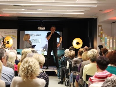 Artur Dziurman z mikrofonem w ręku przemawia do publiczności zgromadzonej w bibliotece. Obok niego na scenie stoi prowadząca spotkanie kobieta w szarym żakiecie, trzymająca dokumenty. W tle wyświetlany jest slajd z nazwiskiem aktora