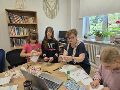 Kobieta w okularach oraz trzy dziewczynki siedzą przy drewnianym stole, wspólnie składając białe arkusze papieru. Na stole widać otwarty laptop, nożyczki, klej w sztyfcie oraz przybory plastyczne.