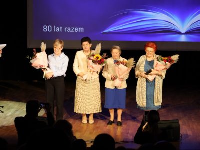 Cztery kobiety w eleganckich strojach stoją na środku sceny, trzymając w dłoniach duże bukiety kwiatów z ozdobną trawą pampasową. Po lewej stronie widać fragment postaci prowadzącej wydarzenie przy mikrofonie.