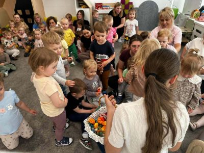 Kadrowane od góry ujęcie w sali zabaw. Na pierwszym planie widać tył głowy i ramiona kobiety trzymającej biały, wiklinowy koszyk wypełniony wielobarwnymi maskotkami z włóczki. Obok stoi mała dziewczynka w jasnej bluzce, uważnie patrząc na zawartość kosza. W tle na dywanie siedzi i bawi się grupa maluchów pod opieką dorosłych kobiet