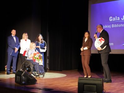 Mężczyzna przemawia do mikrofonu, trzymając ozdobny zegar z napisem „Jubileusz 80-lecia” ozdobiony czerwoną kokardą. Obok stoi kobieta w brązowym garniturze oraz dyrektor biblioteki trzymająca bukiet kwiatów.
