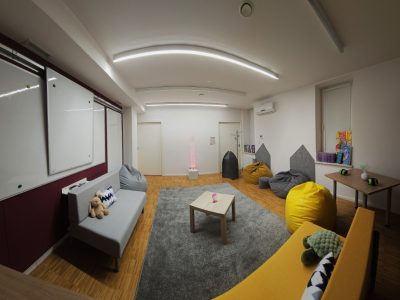 Panorama sali wyciszeń z perspektywy narożnika. Widoczne białe tablice suchościeralne, szara sofa, kolorowe pufy rozłożone na drewnianej podłodze oraz duży szary dywan w centralnej części. W oddali widać podświetloną kolumnę wodną