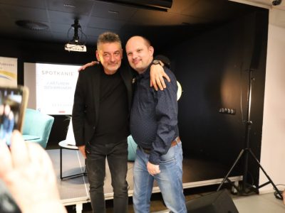 Artur Dziurman pozuje na scenie do zdjęcia z uśmiechniętym mężczyzną w niebieskiej koszuli w kropki i dżinsach, obejmując go ramieniem. Na pierwszym planie z lewej strony widoczna jest dłoń innej osoby robiącej im zdjęcie smartfonem
