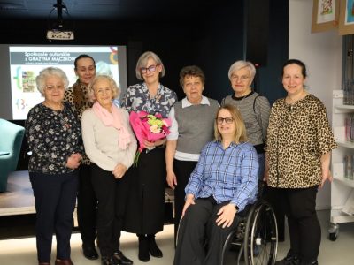 Zdjęcie grupowe po spotkaniu autorskim. Grażyna Mączkowska, trzymająca bukiet kwiatów, stoi w otoczeniu siedmiu czytelniczek w przestrzeni biblioteki