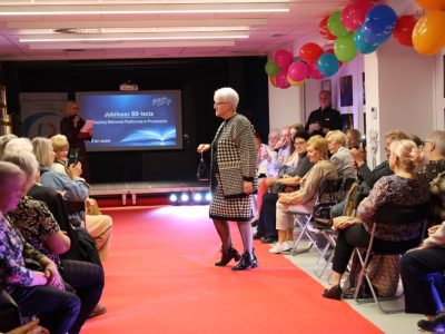 Kobieta w dopasowanej spódnicy i żakiecie we wzór czarno-białej jodełki prezentuje stylizację, idąc po czerwonym dywanie wzdłuż rzędów widzów