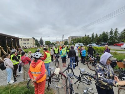 Duża grupa rowerzystów w różnym wieku zebrała się na wybrukowanym placu obok drewnianej tężni solankowej. Wielu uczestników ma na sobie odblaskowe kamizelki oraz kaski ochronne. W tle widoczne są zaparkowane samochody, miejska zabudowa oraz pochmurne niebo