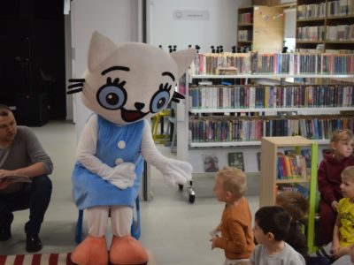 Maskotka Kici Koci siedzi na krześle i wyciąga dłoń w stronę stojącego przed nią chłopca w pomarańczowej bluzce. Obok nich siedzą inne dzieci oraz kuca mężczyzna trzymający otwartą książkę. W tle znajdują się białe regały z literaturą dziecięcą