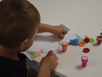 Widok zza ramienia małego chłopca siedzącego przy białym stole, który stawia kolorowe pieczątki na karcie pracy z ilustracją jelenia. Po prawej stronie leżą porozrzucane plastikowe stempelki oraz ich zatyczki