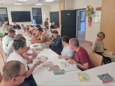 Szerokie ujęcie jaśniejszej sali bibliotecznej lub warsztatowej. Przy dwóch połączonych, białych stołach siedzi kilkunastu uczestników spotkania. Przed nimi rozłożone są liczne książki. Część osób rozmawia ze sobą, inne przeglądają strony. Po prawej stronie przy stole siedzi osoba na wózku inwalidzkim