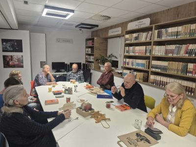 Grupa dorosłych siedząca przy stole w bibliotece podczas spotkania Dyskusyjnego Klubu Książki. Na stole leżą książki, napoje oraz materiały promocyjne z napisem „Wataha”. W tle widać regały pełne książek