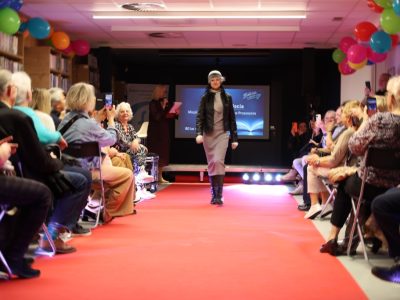Modelka prezentuje stylizację składającą się z czarnej skórzanej kurtki, szarej sukienki dzianinowej, szarej czapki i szala. Idzie sprężystym krokiem po czerwonym dywanie, podczas gdy widzowie robią zdjęcia smartfonami