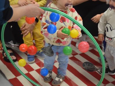 Maluchy uczestniczące w zabawie ruchowej. Dziecko w jasnej bluzie trzyma plastikowe piłeczki przed zielonym obręczem hula-hoop, na który naklejone są kolorowe elementy i ilustracje owadów