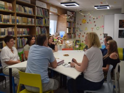 Kadrowane z boku ujęcie uczestników spotkania siedzących wokół stołu w strefie dziecięcej biblioteki. Na pierwszym planie widoczny jest mężczyzna w błękitnej koszulce (tyłem) oraz kobieta w białym t-shircie trzymająca długopis. W tle, na jasnej ścianie ozdobionej papierowymi dekoracjami, widnieje duży, kolorowy napis „LATO”. Obok okna, na półkach i szafkach, stoją liczne pluszowe misie