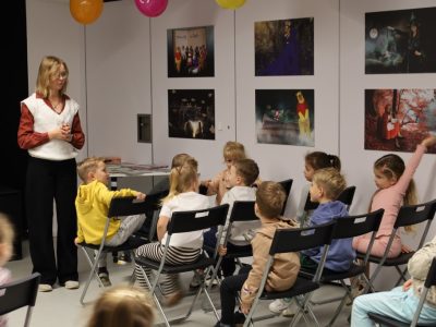 Autorka w białej kamizelce i ciemnych spodniach rozmawia z dziećmi siedzącymi w rzędach na czarnych krzesłach. Na ścianie za dziećmi wiszą oprawione zdjęcia z motywami bajkowymi