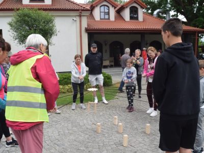 Uczestnicy pikniku biorą udział w zabawie integracyjnej na wybrukowanym dziedzińcu przed budynkiem o dachu z czerwonej dachówki. Trzymając w dłoniach naprężone sznurki, wspólnie sterują metalowym chwytakiem zawieszonym na środku, próbując ustawić pionowo drewniane klocki na ziemi