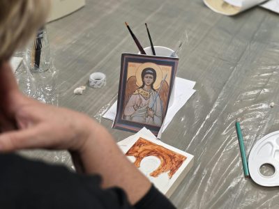Spojrzenie zza pleców uczestniczki warsztatów na stół, na którym leży wzór ikony z Aniołem Stróżem w kubku oraz podkładka z rozpoczętym malunkiem