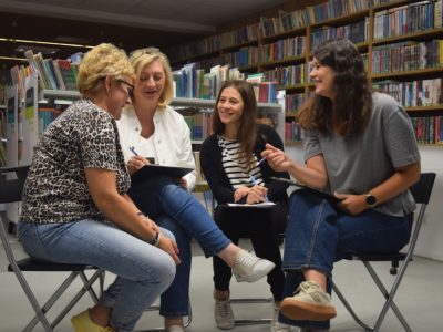 Bliskie ujęcie na cztery uczestniczki warsztatów siedzące naprzeciwko siebie w przestrzeni biblioteki. Kobiety rozmawiają, uśmiechają się i notują wnioski w podkładkach biurowych