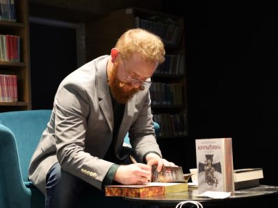 Zbliżenie na Krzysztofa Piersę pochylonego nad stolikiem i składającego autograf w książce. Obok stoi egzemplarz jego powieści „Konfraternia”
