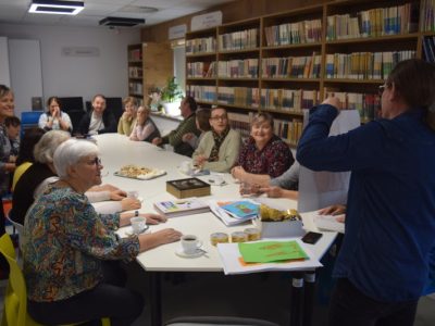 Spotkanie podsumowujące warsztaty przy połączonych białych stołach w bibliotece. Mężczyzna w niebieskiej koszuli stoi przodem do grupy i unosi białą planszę z gotową pracą malarską. Wokół stołu siedzi kilkanaście osób, rozmawiając i patrząc w jego stronę. Na blacie leżą rolki taśmy klejącej oraz wykonane rysunki
