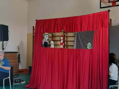 W centralnej części zdjęcia widać czerwoną kotarę teatralną. W jej głównej części widać postawi Czerwonego Kapturka, Babci oraz Wilka. Po lewej stronie konstrukcji siedzi kobieta, która trzyma w rękach scenariusz przedstawienia. Jest narratorem