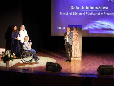 Mężczyzna w eleganckiej marynarce w kratkę przemawia do mikrofonu na środku sceny, stojąc obok stojącego portretu historycznego i dyrektor bibliotek