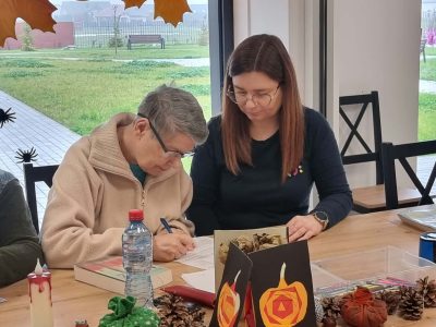 Kobieta w okularach po lewej stronie pisze na kartce papieru przy drewnianym stole, obok siedzi opiekunka. Na stole stoją jesienne dekoracje z papierowych dyń i szyszek