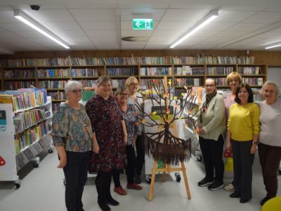 Grupa ośmiu uśmiechniętych kobiet stoi w rzędzie w bibliotece, pozując do zdjęcia wokół gotowego rękodzieła. Na drewnianej sztaludze pośrodku umieszczono okrągłą makramę z brązowych sznurków przypominającą drzewo, którego gałęzie udekorowano papierowymi, kolorowymi listkami. W tle widać białe regały z książkami dziecięcymi i napisami „Poziom I”