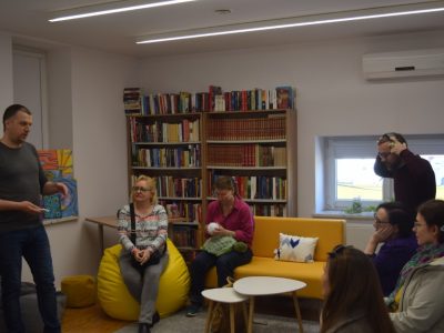 Mężczyzna stoi i przemawia do grupy osób zgromadzonych w bibliotecznym pokoju wyciszeń. Uczestnicy siedzą na żółtej sofie oraz żółtej pufie typu bean bag. W tle widać regały z książkami