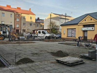 Teren budowy przed parterowym, żółtym budynkiem z napisem „Dom Harcerza”. Szeroki plac ma wyrównane, piaszczyste podłoże przygotowane pod układanie kostki. Po lewej stronie widać fragment żółtej koparki, a na placu pracują robotnicy – jeden z nich obsługuje zagęszczarkę spalinową. Na pierwszym planie leży pusta drewniana paleta. W tle stoją kamienice oraz zaparkowane samochody