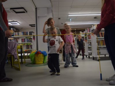 Dzieci biorą udział w zabawie pod nazwą "limbo" w bibliotece. Bibliotekarka i wolontariuszka trzymają rozciągnięty żółty sznur, pod którym przechodzi uśmiechnięty chłopiec w białej koszulce. Pozostałe dzieci czekają w kolejce, obserwując zabawę