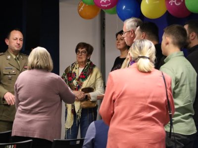 Dariusz Kwiatkowski w mundurowym garniturze stoi po lewej stronie i rozmawia w kuluarach z grupą uczestników wydarzenia. W centrum widać kobietę w barwnej chustce, a nad głowami uczestników wiszą kolorowe balony