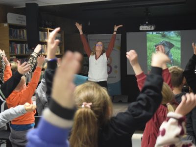 Marta Maj w okularach i białej kamizelce stoi na tle ekranu ze zdjęciem psa i unosi ręce do góry. Siedzące przed nią dzieci również podnoszą dłonie w ramach wspólnej zabawy