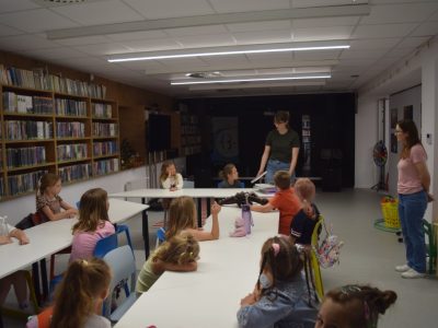 Grupa dzieci siedzi przy białych stołach ustawionych w salce bibliotecznej, słuchając stojącej przed nimi prowadzącej w ciemnozielonej koszulce. Z boku przygląda się im druga opiekunka, a po lewej stronie widać regały pełne książek