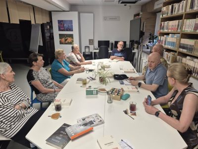 Dyskusja grupy czytelników siedzących przy białym stole w nowoczesnej przestrzeni bibliotecznej. Na blacie stołu, obok filiżanek z napojami, wyeksponowane są okładki różnych książek przygotowanych do rozmowy. W tle, poza regałami z książkami, widoczne są stanowiska komputerowe oraz dekoracyjne grafiki na ścianach