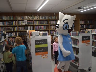Spacer między regałami. Maskotka Kicia Kocia idzie korytarzem bibliotecznym, a za nią i obok niej między białymi regałami przemykają dzieci. Widoczne są oznaczenia poziomów książek dla dzieci
