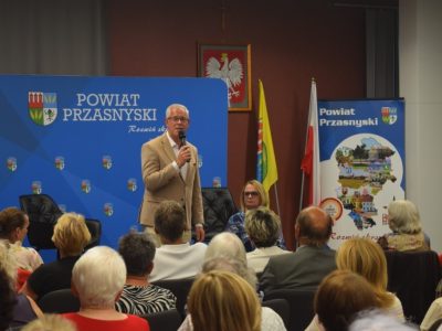 Mężczyzna w beżowej marynarce przemawia do mikrofonu przed widownią. W tle niebieska ścianka z logo Powiatu Przasnyskiego oraz polskie flagi