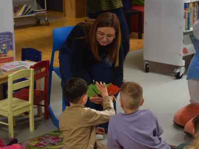 Bibliotekarka pochyla się w stronę dzieci siedzących na dywanie, pokazując im kolorowe wycinanki w kształcie głowy kota (czerwony, zielony, żółty). Kobieta uśmiecha się do chłopca, który wyciąga rękę w stronę kolorowych pomocy dydaktycznych. Obok widać mały stolik z książeczkami z serii o Kici Koci