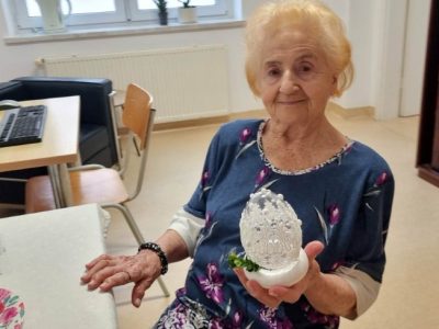 W centralnej części zdjęcia znajduje się uśmiechnięta kobieta, która trzyma prezentuje w dłoni gotową ozdobę wielkanocną. Za nią po lewej stronie stoi komputer dla użytkowników. Po prawej stronie zdjęcia znajduje się brązowa witryna.