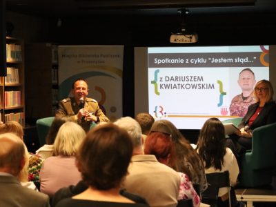 Na scenie siedzą dwie osoby: po lewej generał Dariusz Kwiatkowski w garniturze wojskowym z mikrofonem w ręku, po prawej prowadząca spotkanie kobieta. W tle widać duży ekran projektora z napisem „Spotkanie z cyklu Jestem stąd… z Dariuszem Kwiatkowskim” oraz jego zdjęciem. Przed sceną siedzi widownia widoczna od tyłu