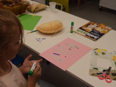 Dziewczynka w okularach siedzi przy białym stole, trzymając w dłoni klej w sztyfcie. Przed nią leży różowy arkusz papieru z naklejonymi elementami, zielona kartka, nożyczki, ozdobna poduszka oraz czasopismo „Świerszczyk”