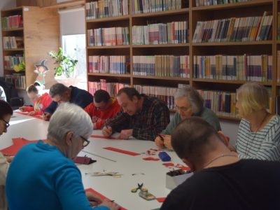 Bliskie ujęcie na osoby siedzące przy stole przed regałami z książkami. Uczestnicy za pomocą ołówków i linijek odmierzają i odrysowują linie na czerwonych arkuszach papieru, przygotowując elementy do wycięcia
