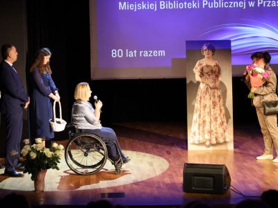 Kobieta w jasnym żakiecie i z przewieszoną torebką przemawia do mikrofonu, trzymając w dłoniach bukiet kwiatów skierowany w stronę dyrektor
